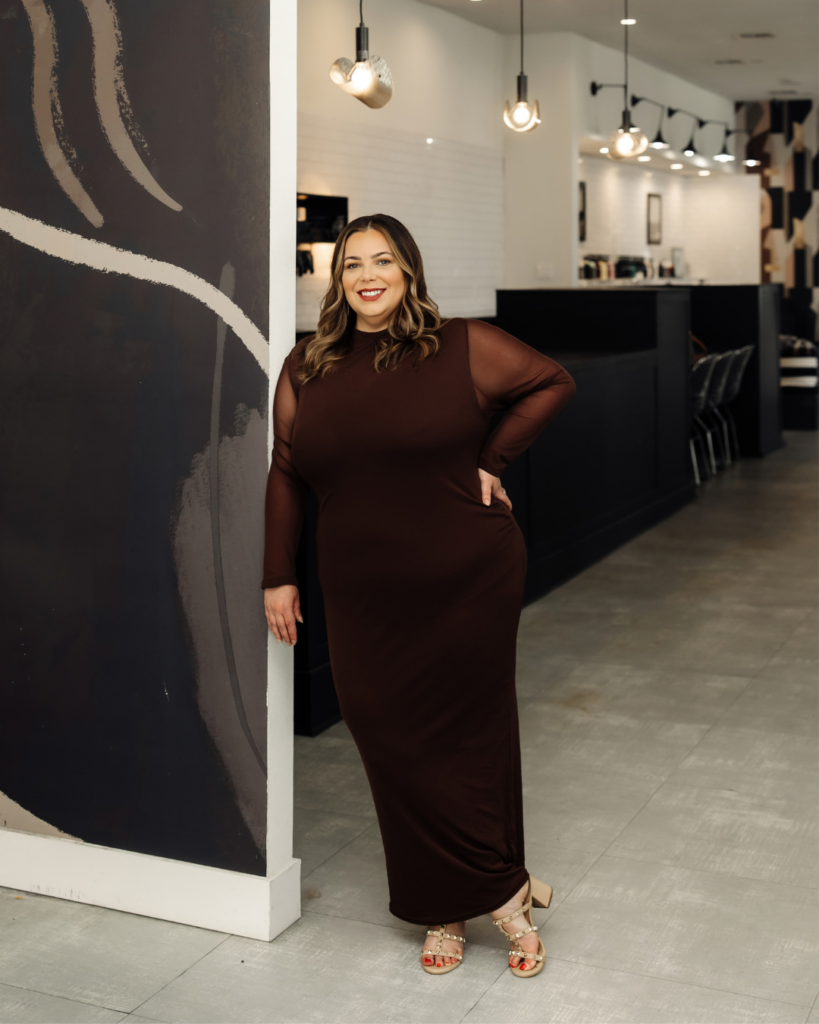 Portrait of stylist Heather Fogarty standing elegantly in a brown long-sleeved dress inside the salon.