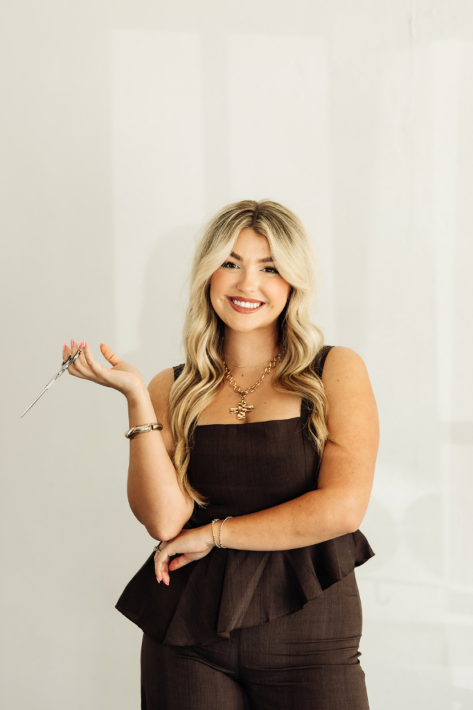Portrait of blonde stylist Kansas Hebert holding hair shears with a warm smile.