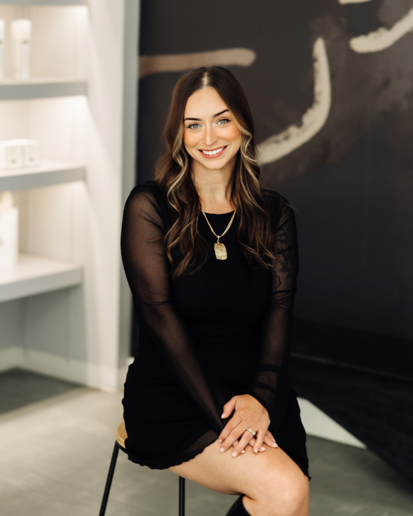 Portrait of brunette stylist Kayla Moore with face-framing highlights sitting on a stool in the salon.